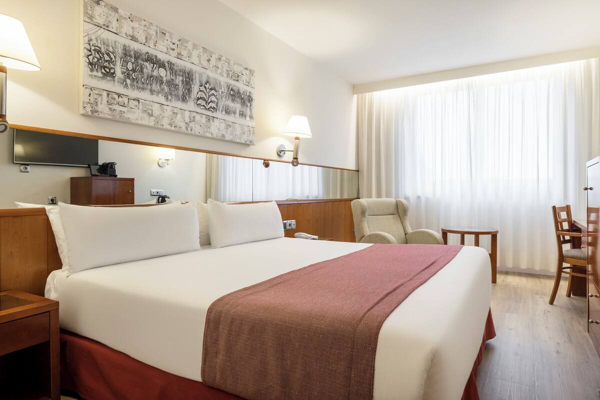 A hotel room with a large bed, two bedside lamps, a flat-screen TV, a wooden nightstand, a desk with a chair, and a large window with curtains.