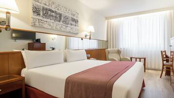 A hotel room with a large bed, two bedside lamps, a flat-screen TV, a wooden nightstand, a desk with a chair, and a large window with curtains.