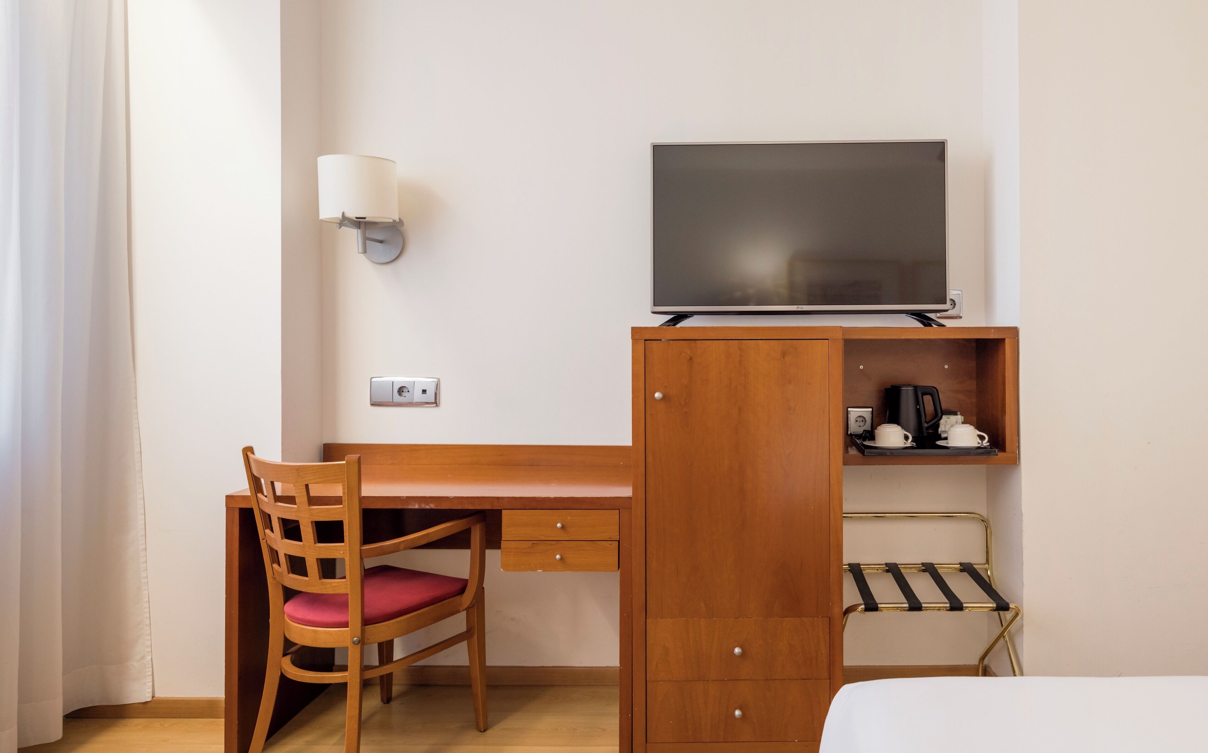 Ett hotellrum med ett skrivbord i trä, en tv, en stol med en röd kudde och en säng.