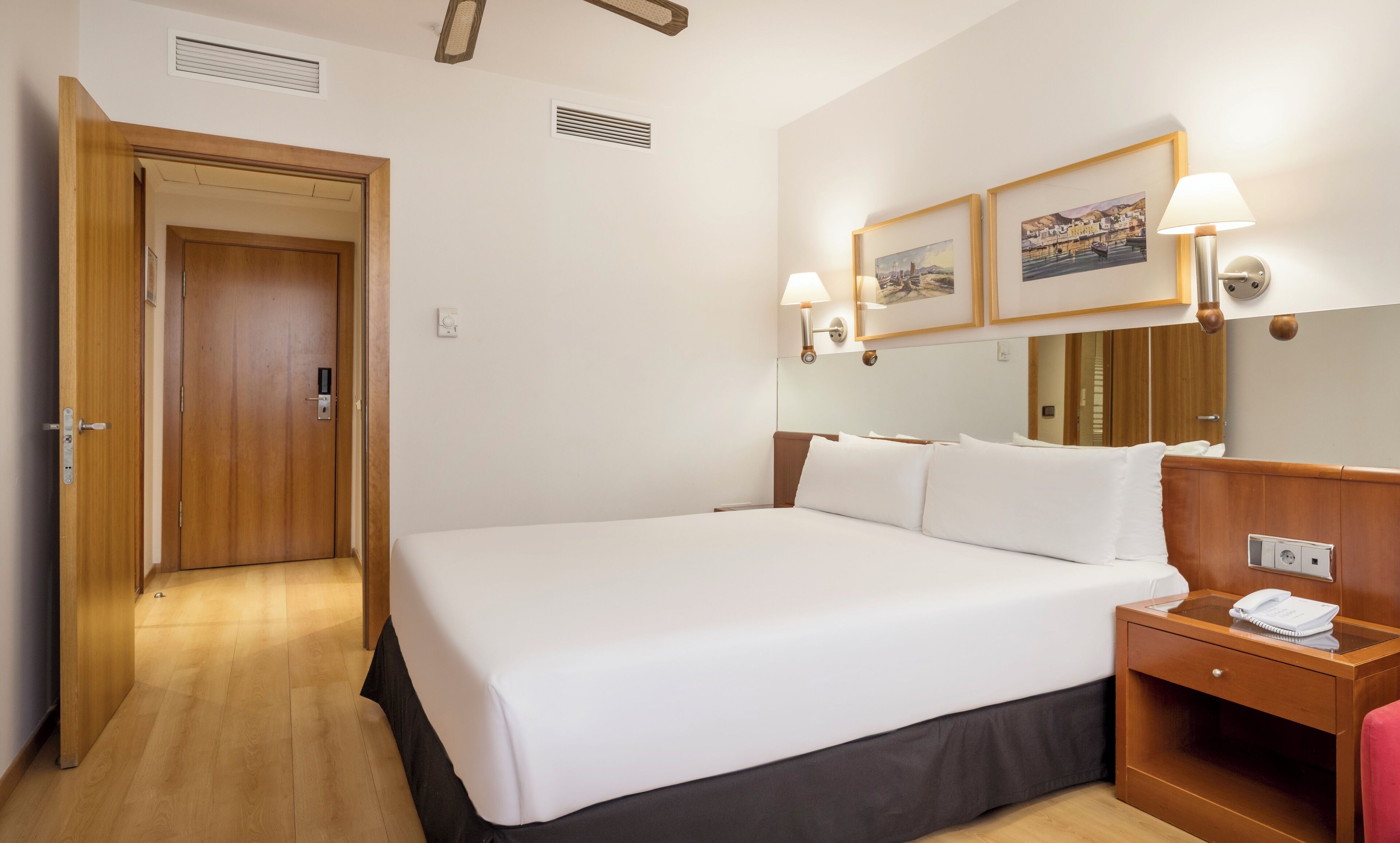 Une chambre d’hôtel avec un grand lit, une tête de lit en bois, des tables de chevet et une lampe fixée au mur.