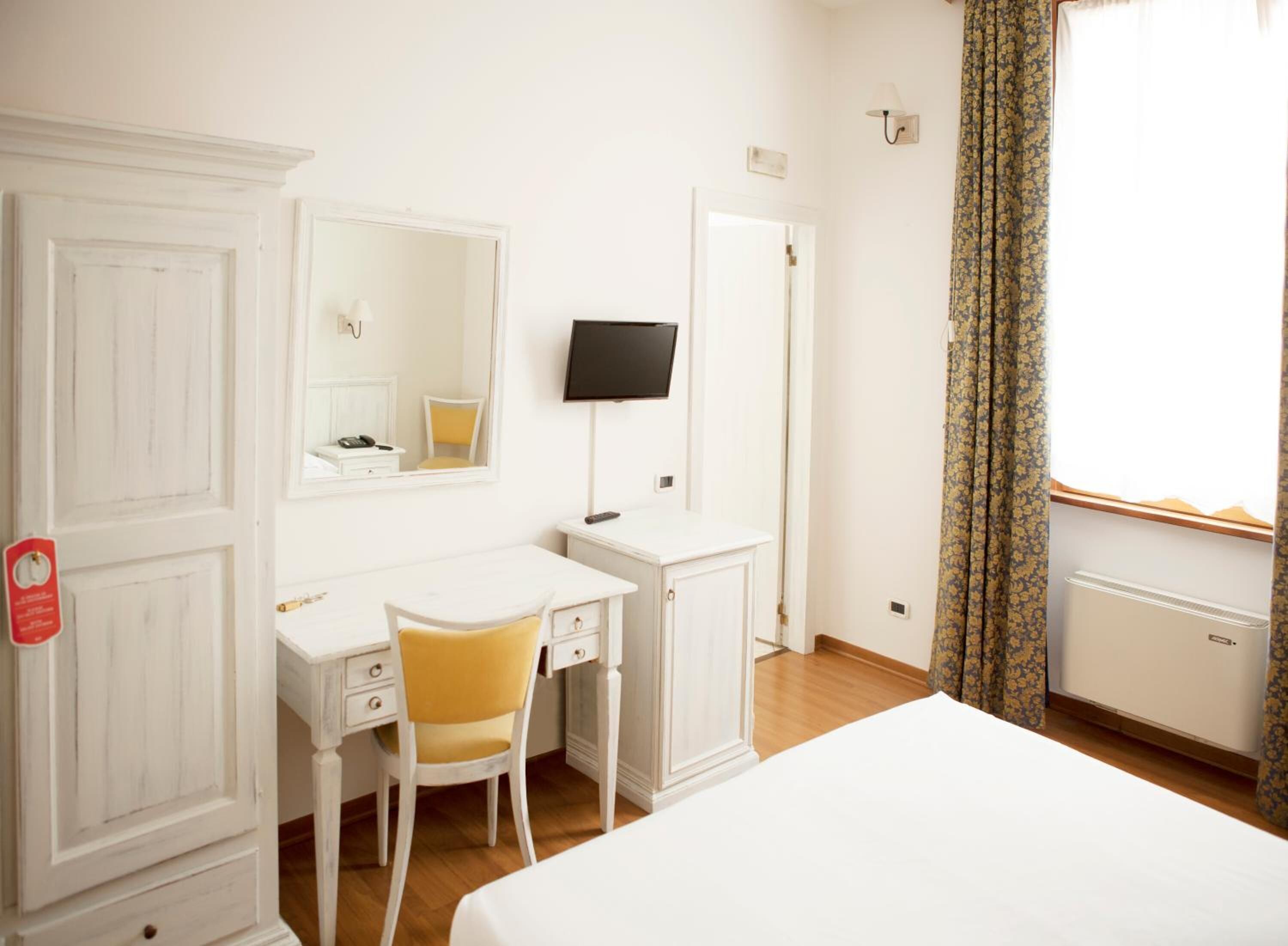 A hotel room with a white desk, a chair, a bed, a television, and a window with curtains.