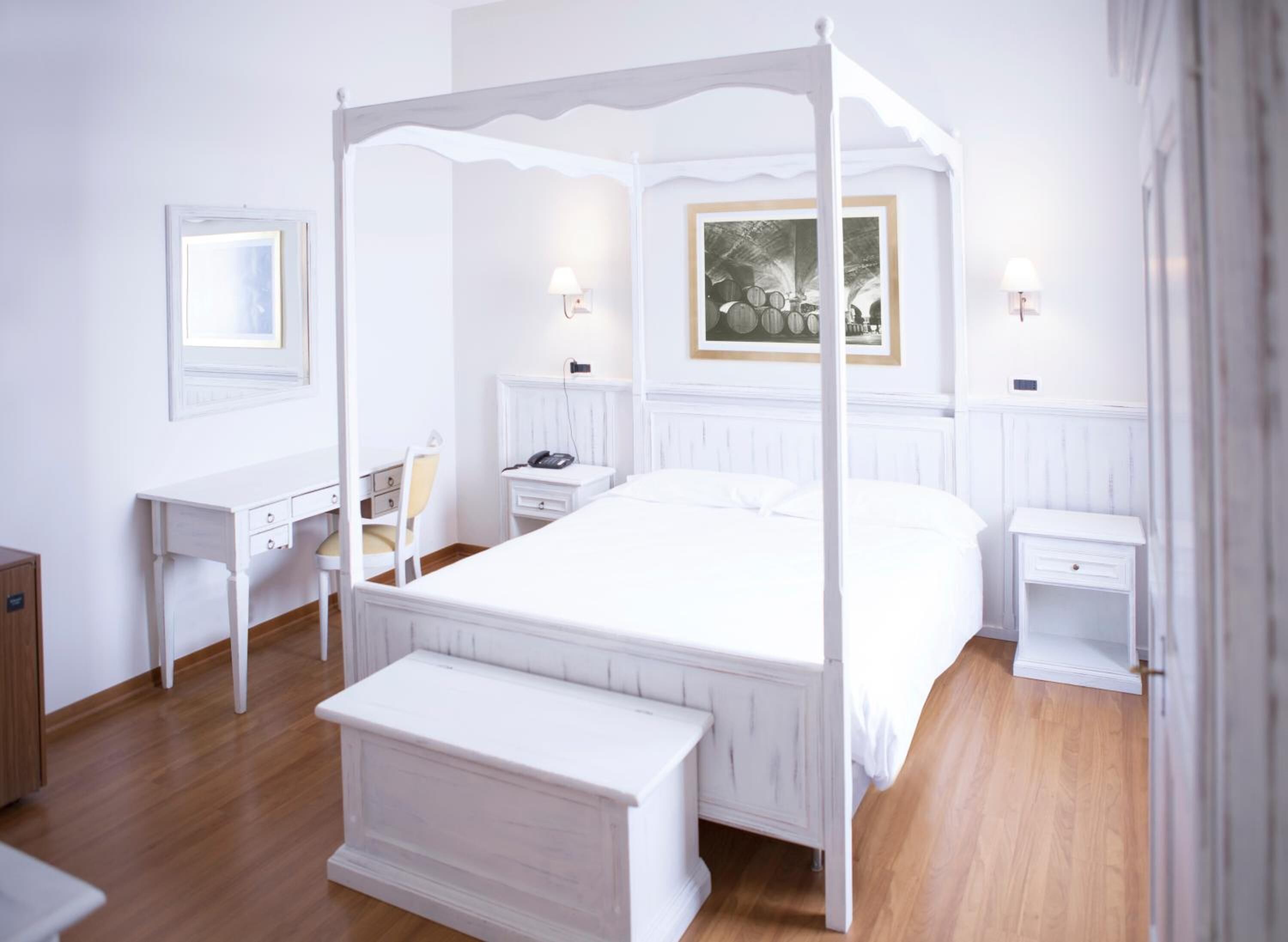 A four-poster bed with a white canopy, a desk with a chair, a bedside table with a lamp, and a framed picture on the wall.
