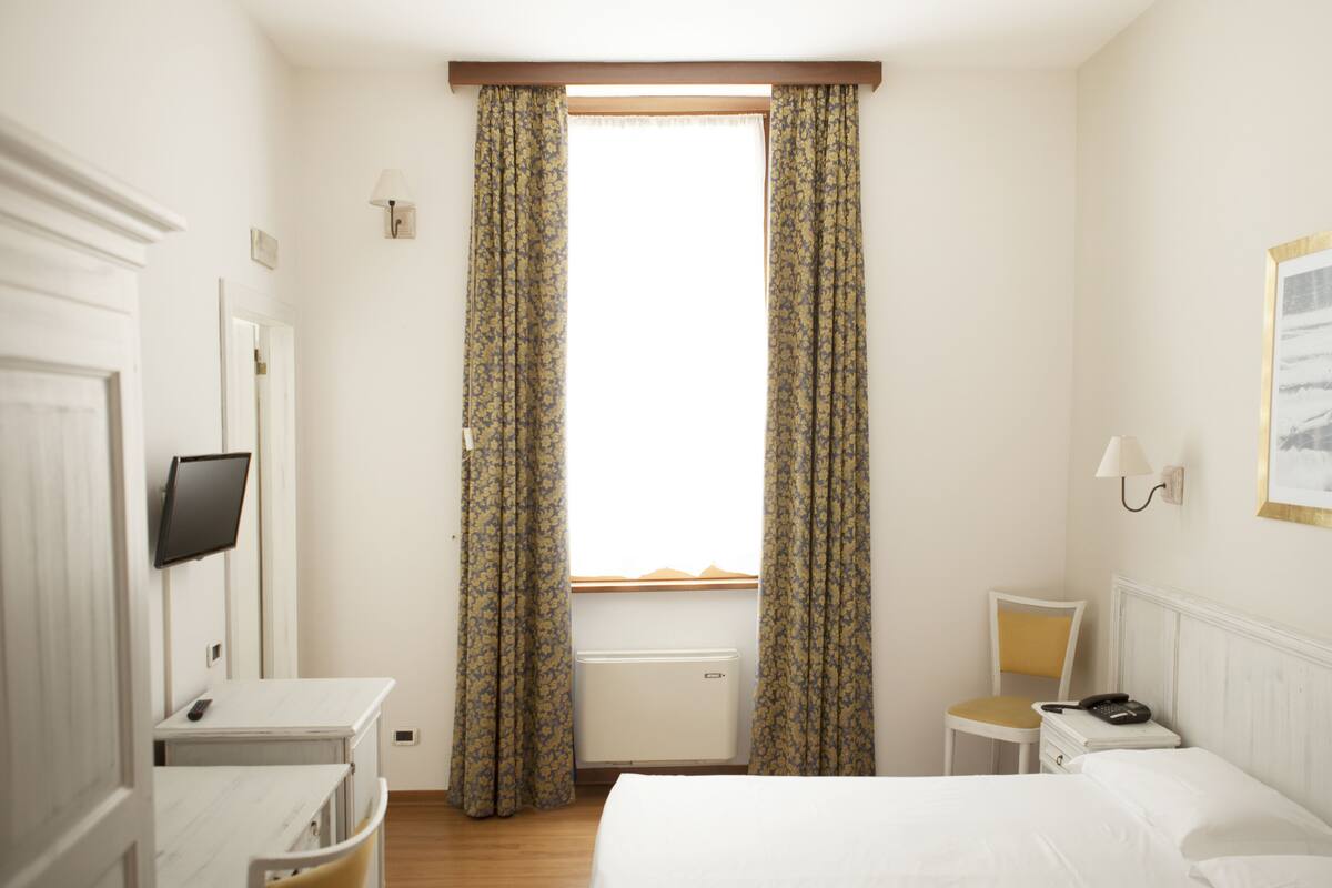 A hotel room with a bed, a desk, a chair, a bedside table with a phone, and a TV mounted on the wall.