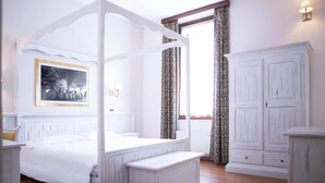 Una cama con dosel, cabecera, mesita de noche, banco y armario en una habitación con un ventanal y cortinas.