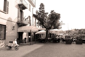 A vintage black and white photo of a cobblestone street with a parked scooter, outdoor tables and chairs under umbrellas, and a building labeled "HOTEL SEBINO".