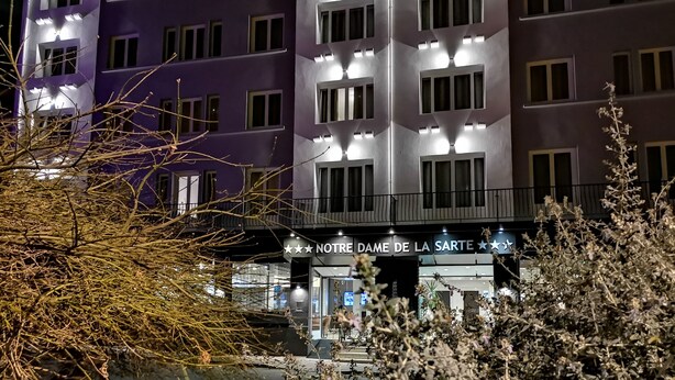A hotel facade with a sign reading "NOTRE DAME DE LA SARTRE" and multiple illuminated windows.