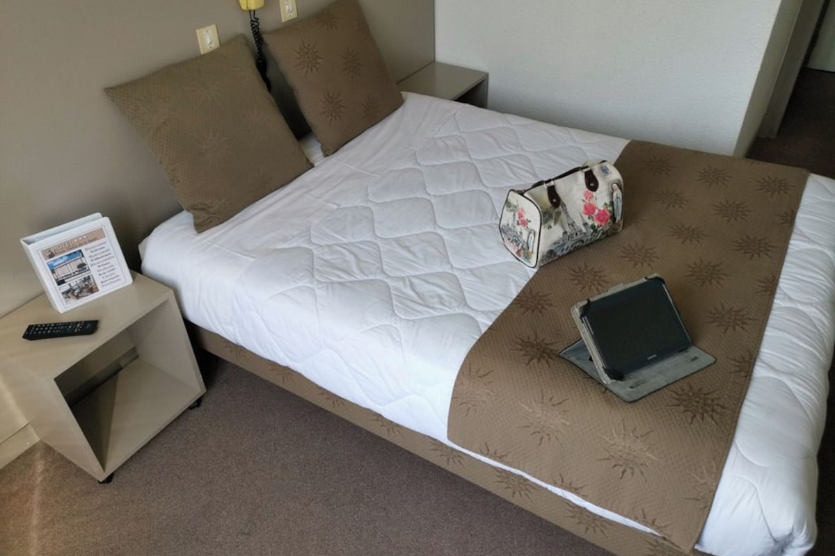 A neatly made bed with a quilted cover, a bedside table with a remote control, a floral-patterned bag, and a laptop on the bed.