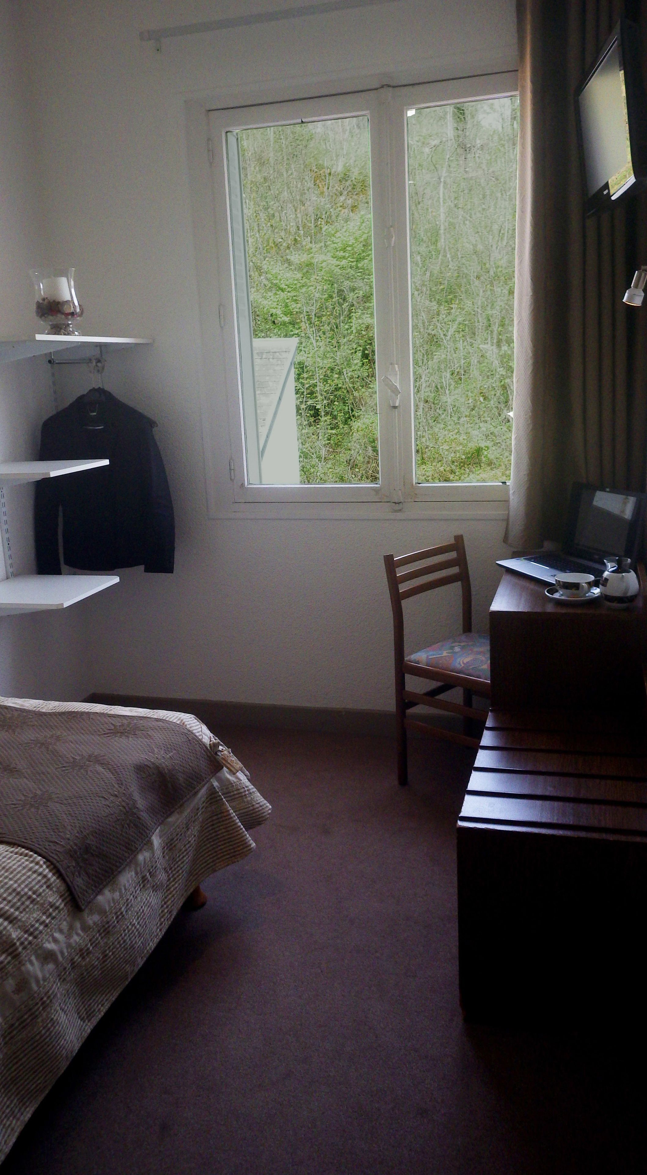 A bedroom with a bed, a chair, a desk with a laptop, a window with a view of greenery, and a shelf with a jacket and a glass.
