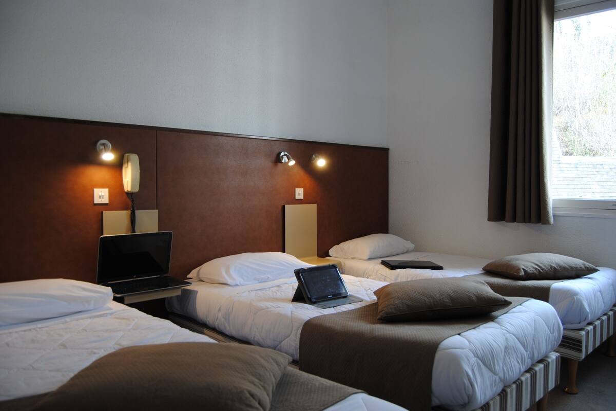 A hotel room with three beds, a laptop on one bed, and a bedside table with a lamp.