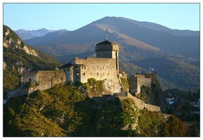 A historic stone castle on a hill with a flag, surrounded by mountains. - Adonis Lourdes (Lourdes)