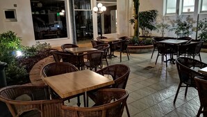 An outdoor seating area with wicker furniture and a tiled floor, surrounded by plants and a view of the interior.