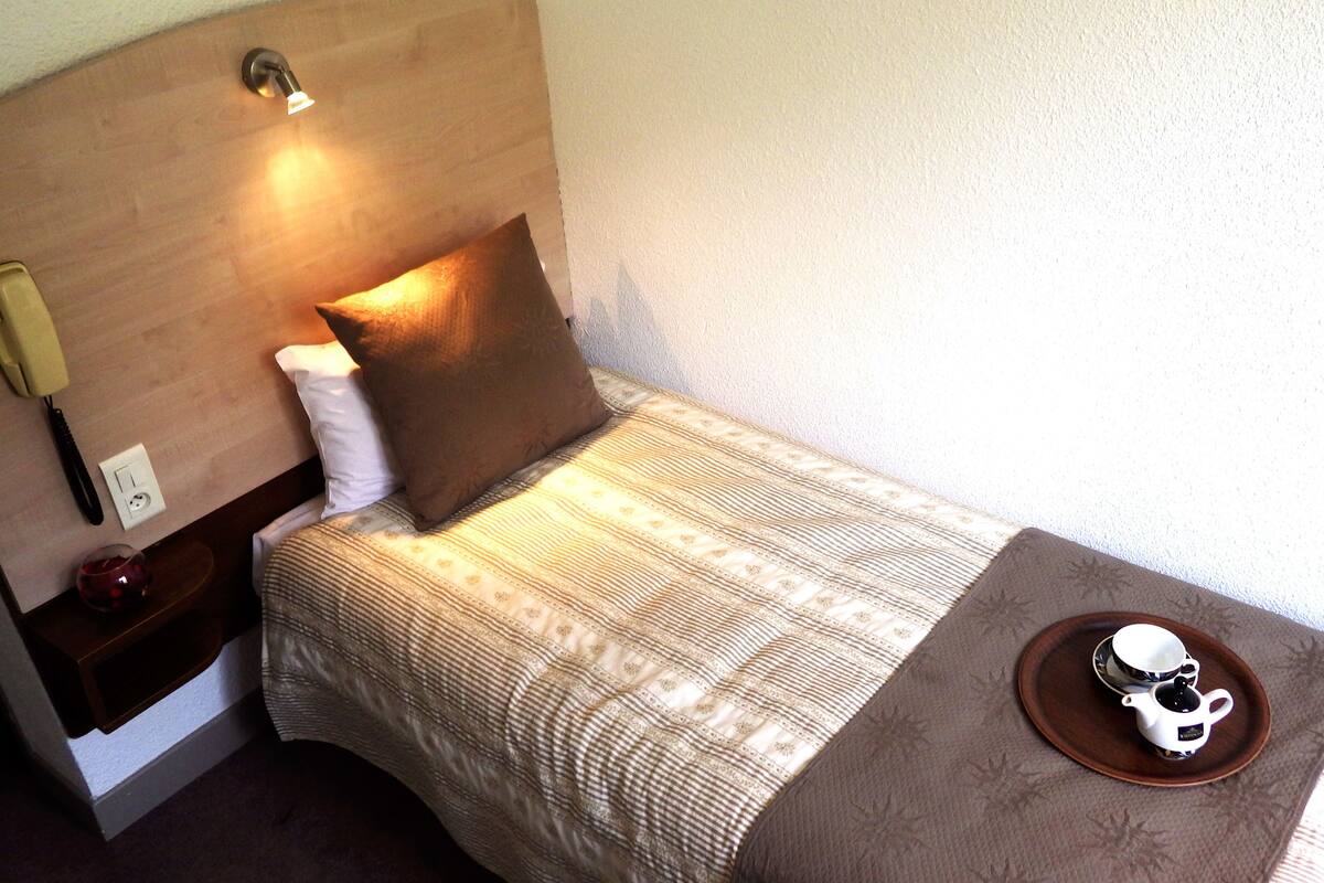 A neatly made bed with a patterned bedspread, a pillow, a bedside lamp, a bedside phone, and a tray with a teapot and cup.