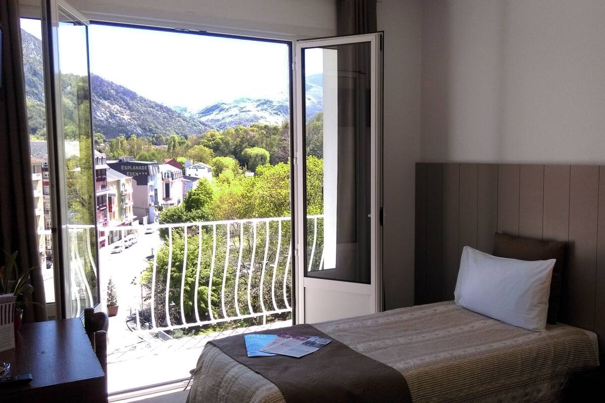 A hotel room with a bed, a bedside telephone, and a view of a balcony with a railing.