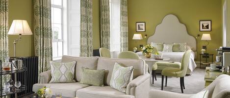 A hotel room with a large bed, a sofa, a coffee table, and a dining table. There are curtains, a chandelier, and a picture on the wall.