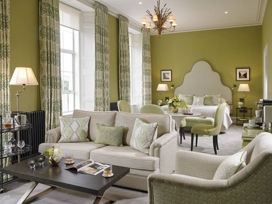 Habitación de hotel con una cama grande, un sofá, una mesa de centro y una mesa de comedor. Hay cortinas, un candelabro y un cuadro en la pared.