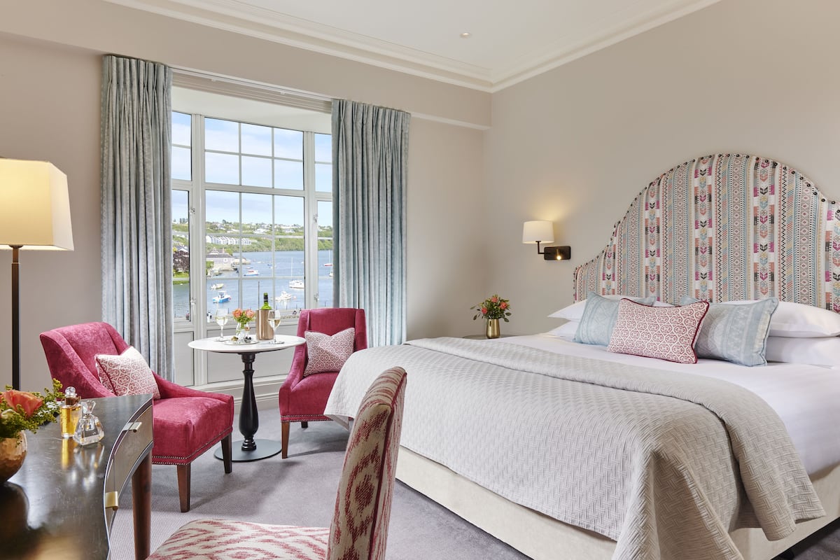 A hotel room with a large bed, a sitting area with a table and chairs, a view of the sea, and a patterned headboard.