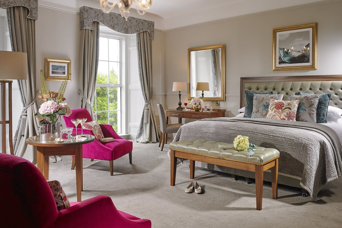 A bedroom with a large bed, a vanity with a mirror, a chandelier, and a view of greenery outside.