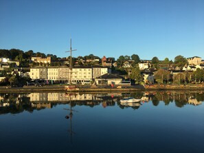 A waterfront hotel with a reflection in the calm water, featuring a prominent building with a flagpole and a boat docked nearby. - Actons Hotel (Kinsale)