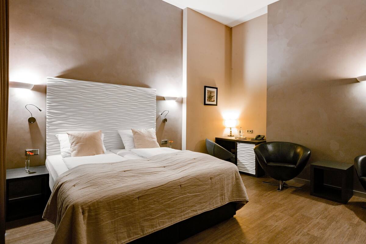 A modern hotel room with a large bed, two bedside tables, a desk, a chair, and a wall-mounted lamp.