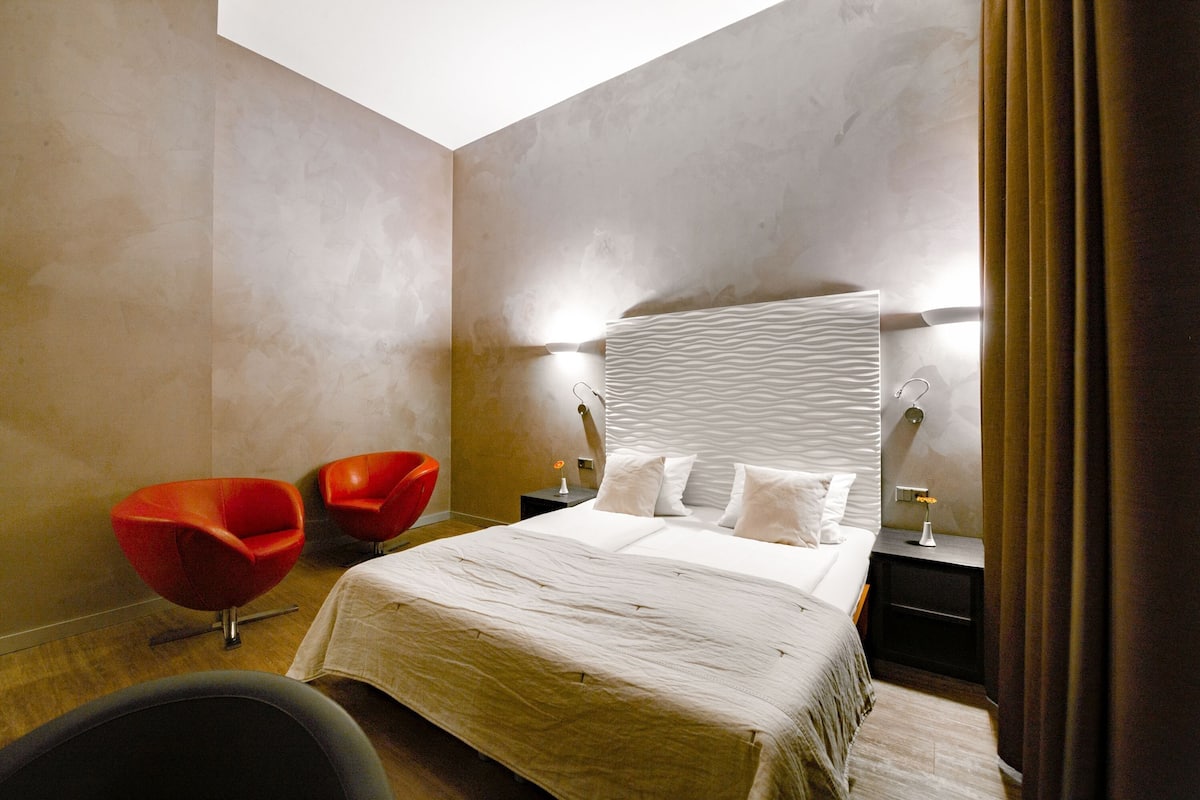 A modern hotel room with a large bed, two red chairs, a nightstand, and a large window.