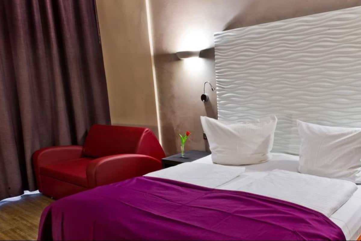 A hotel room with a bed, a red chair, a nightstand, and a wall-mounted lamp.