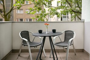 Ein kleiner Balkon mit rundem Tisch und zwei Korbstühlen, einer Vase mit einer Blume sowie Blick auf Gebäude und Bäume im Hintergrund.