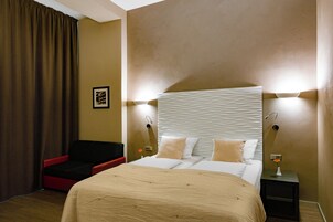 A hotel room with a large bed, a red chair, a nightstand with a lamp, and a wall-mounted light fixture.