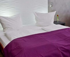 Una cama bien hecha con almohadas blancas y una colcha morada, una mesita de noche con un jarrón de flores, y una pared con textura detrás de la cama.