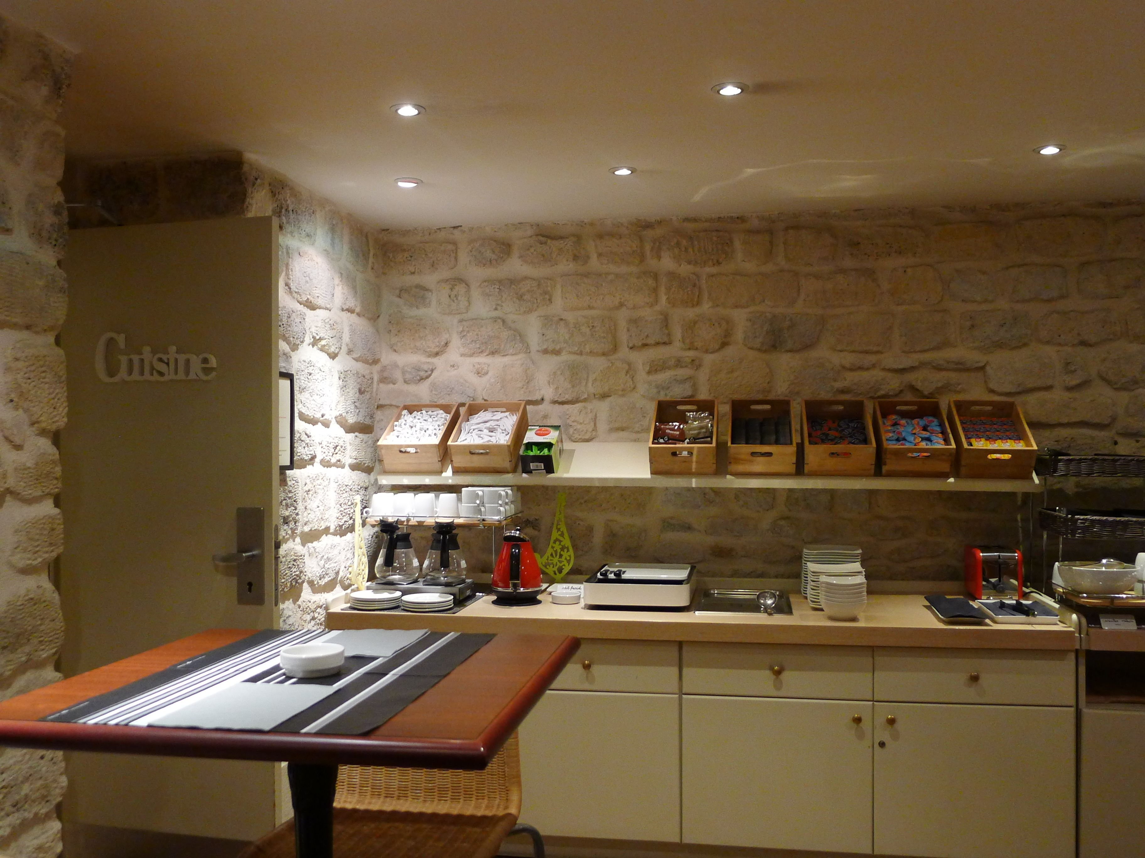 Un coin cuisine avec un mur en pierre, un espace dédié au café et divers produits alimentaires rangés sur des étagères.