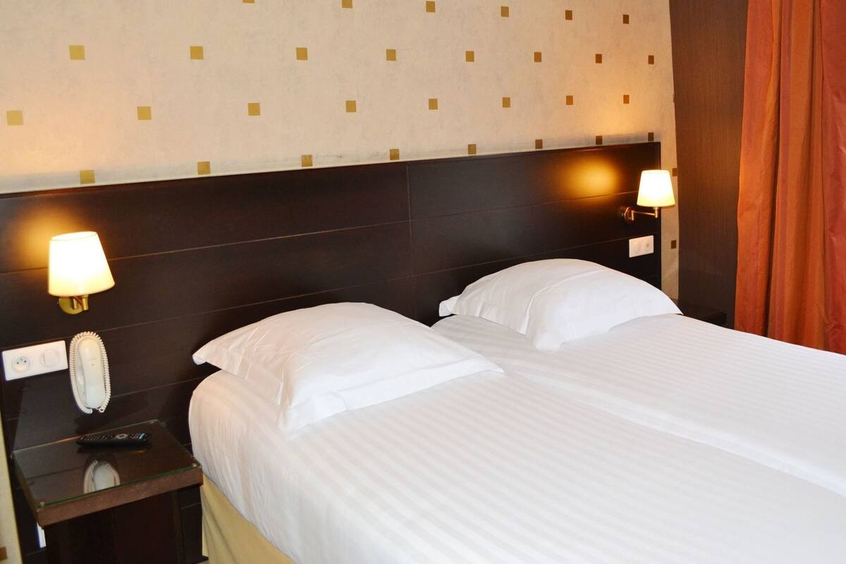 Quarto de hotel com cama, duas luminárias de cabeceira, telefone e janela com cortinas.