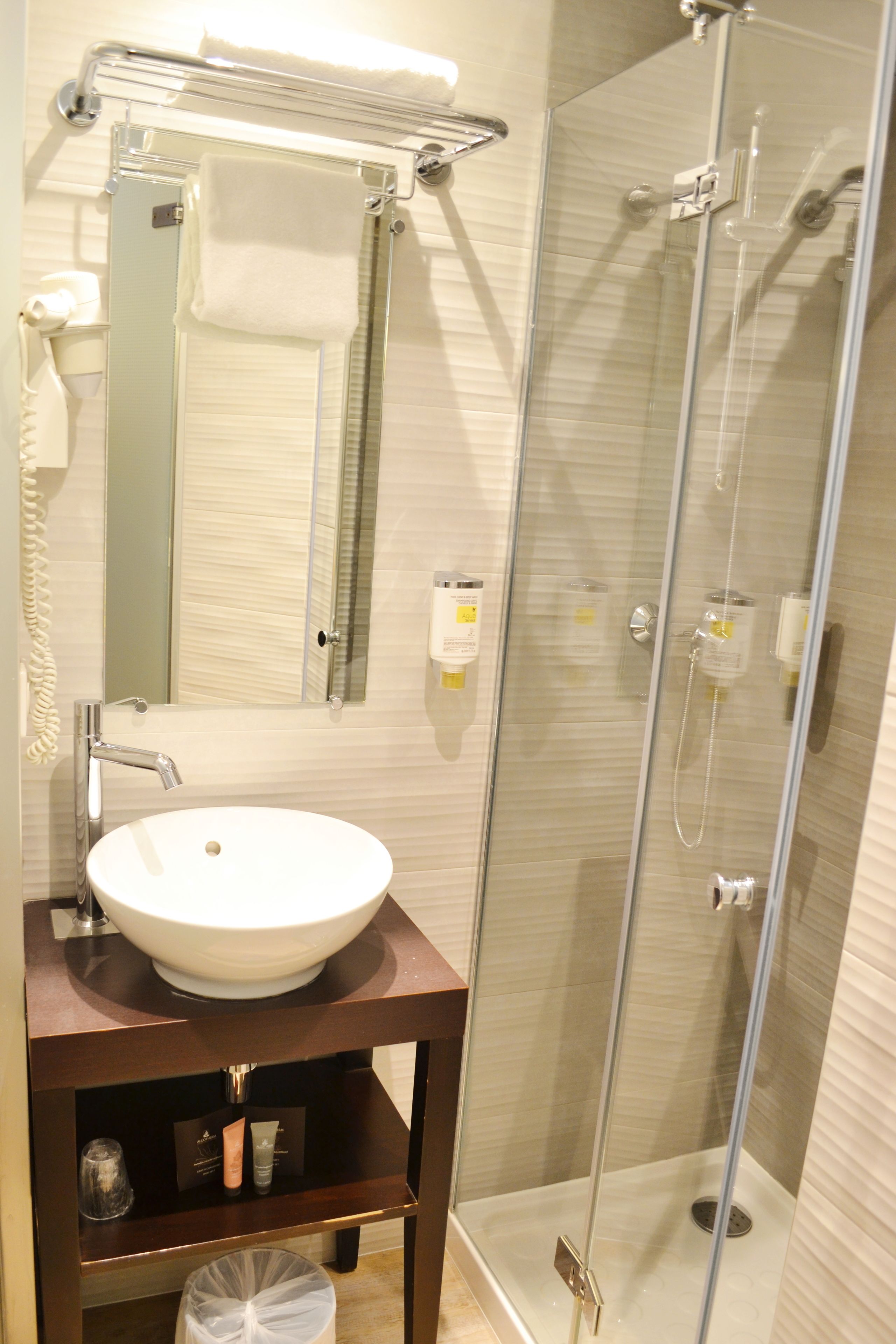 Un baño con un lavabo blanco, un espejo, toallero, ducha con mampara de cristal y un cubo de basura.