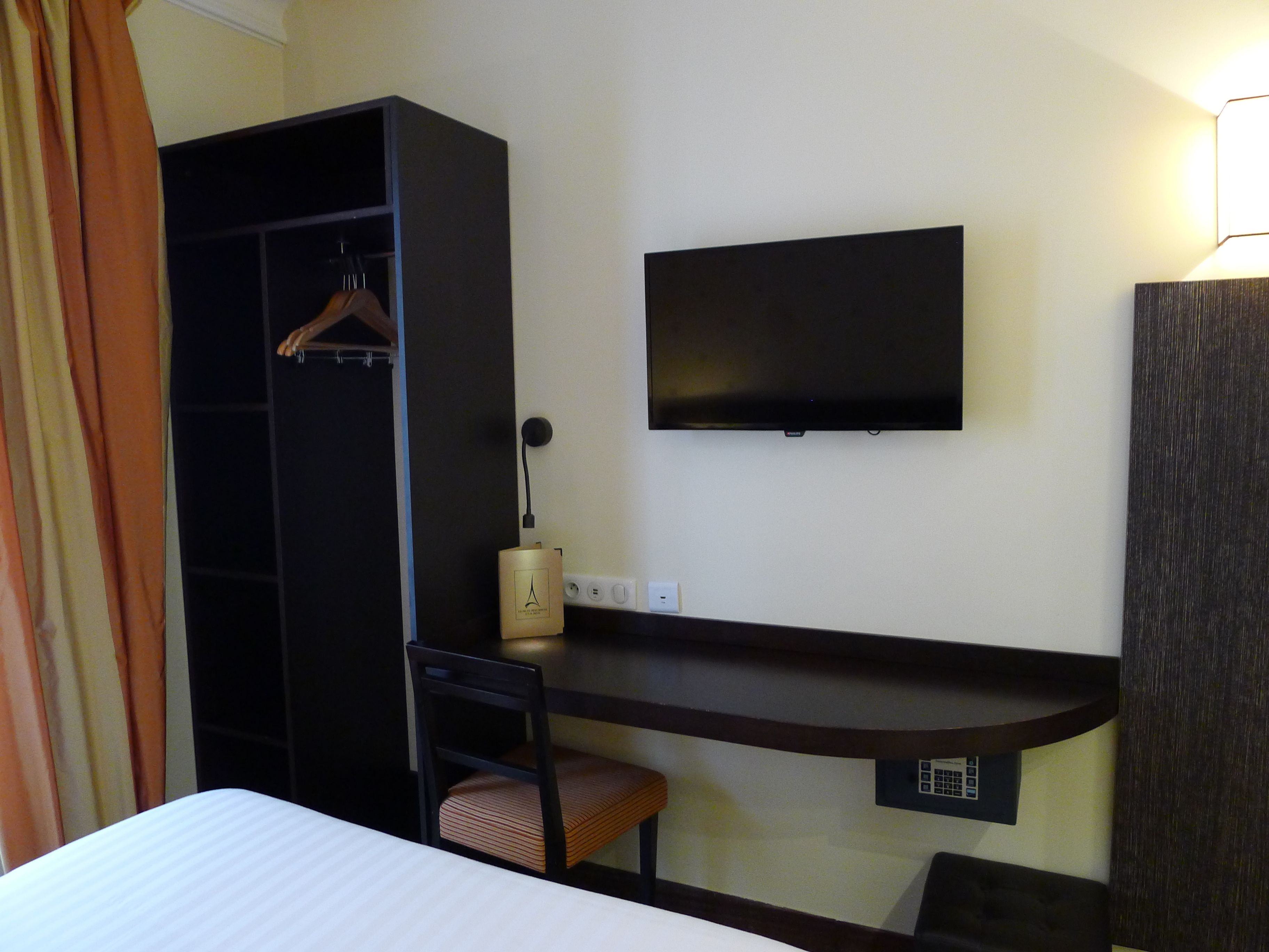A hotel room with a bed, a desk, a chair, a wardrobe, a TV, and a lamp.