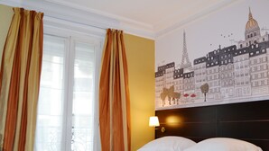 Habitación de hotel con cama, cortinas, un mural de edificios parisinos y una mesita de noche con lámpara.