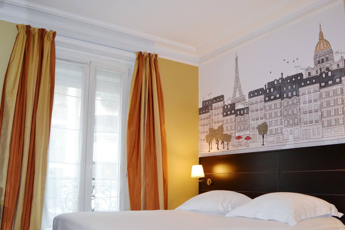 Quarto de hotel com cama, cortinas, um mural na parede com prédios parisienses e uma mesa de cabeceira com abajur.