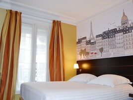 Habitación de hotel con cama, cortinas, un mural de edificios parisinos y una mesita de noche con lámpara.
