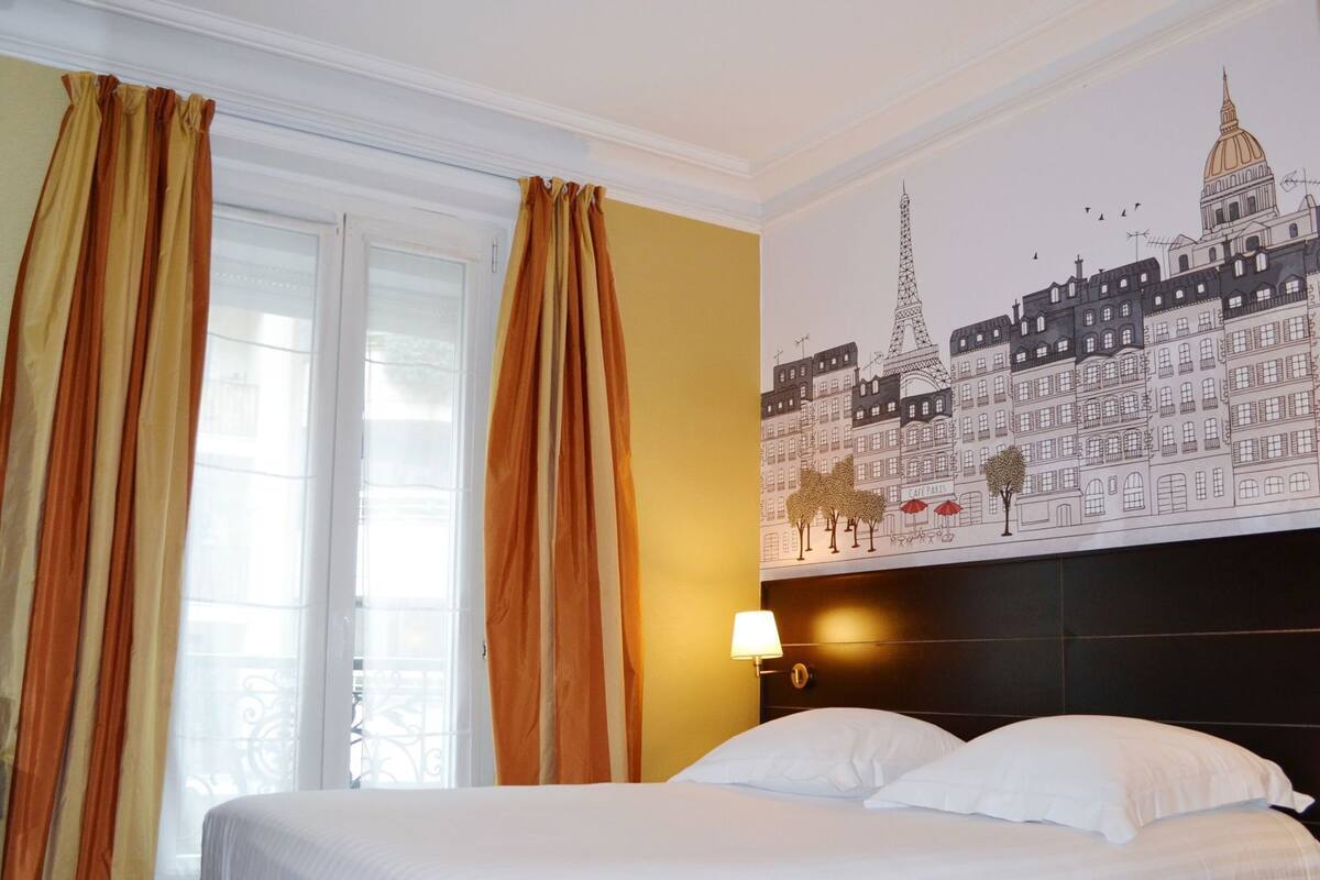 Quarto de hotel com cama, cortinas, painel na parede com prédios parisienses e mesa de cabeceira com abajur.