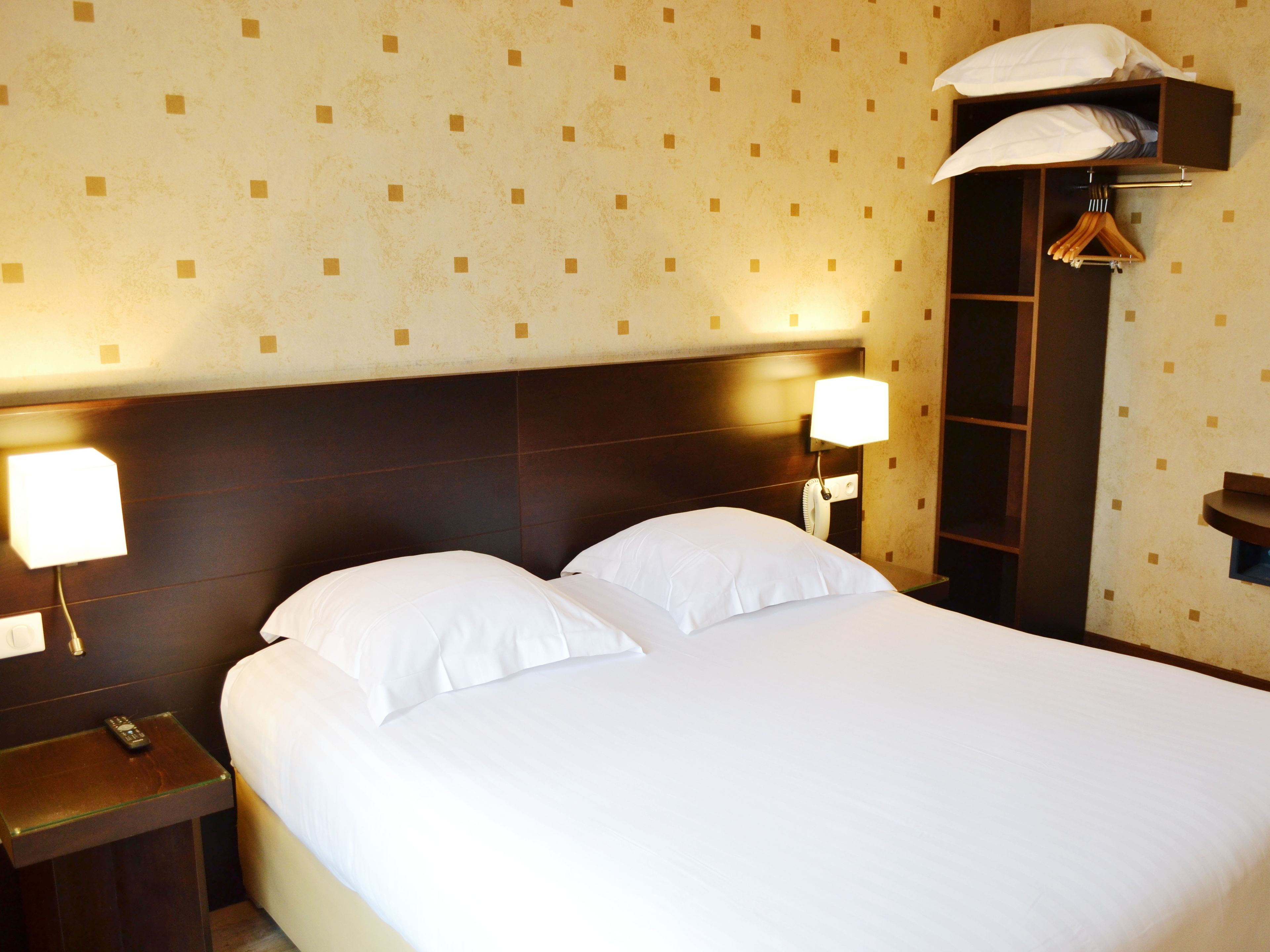 Ein ordentlich bezogenes Bett mit weißen Leinen in einem Hotelzimmer. Es gibt zwei Nachttischlampen, einen Holznachttisch und ein an der Wand montiertes Regal mit einer Kleiderstange.
