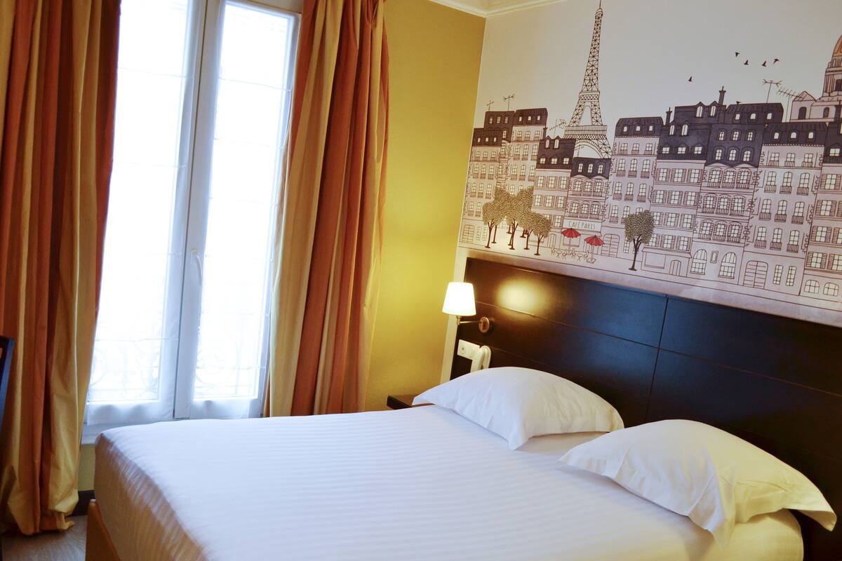 Quarto de hotel com cama, cortinas, um mural na parede, luminária e mesa de cabeceira.