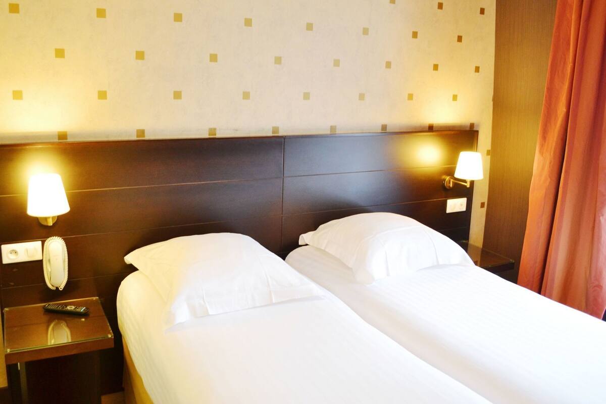 Quarto de hotel com duas camas, cabeceira, abajures, mesa de cabeceira com telefone e mesa de vidro.