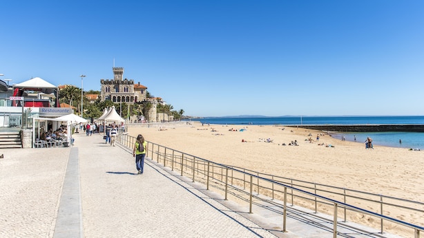 A beachside promenade with a historic building, a restaurant, and a clear blue sky.