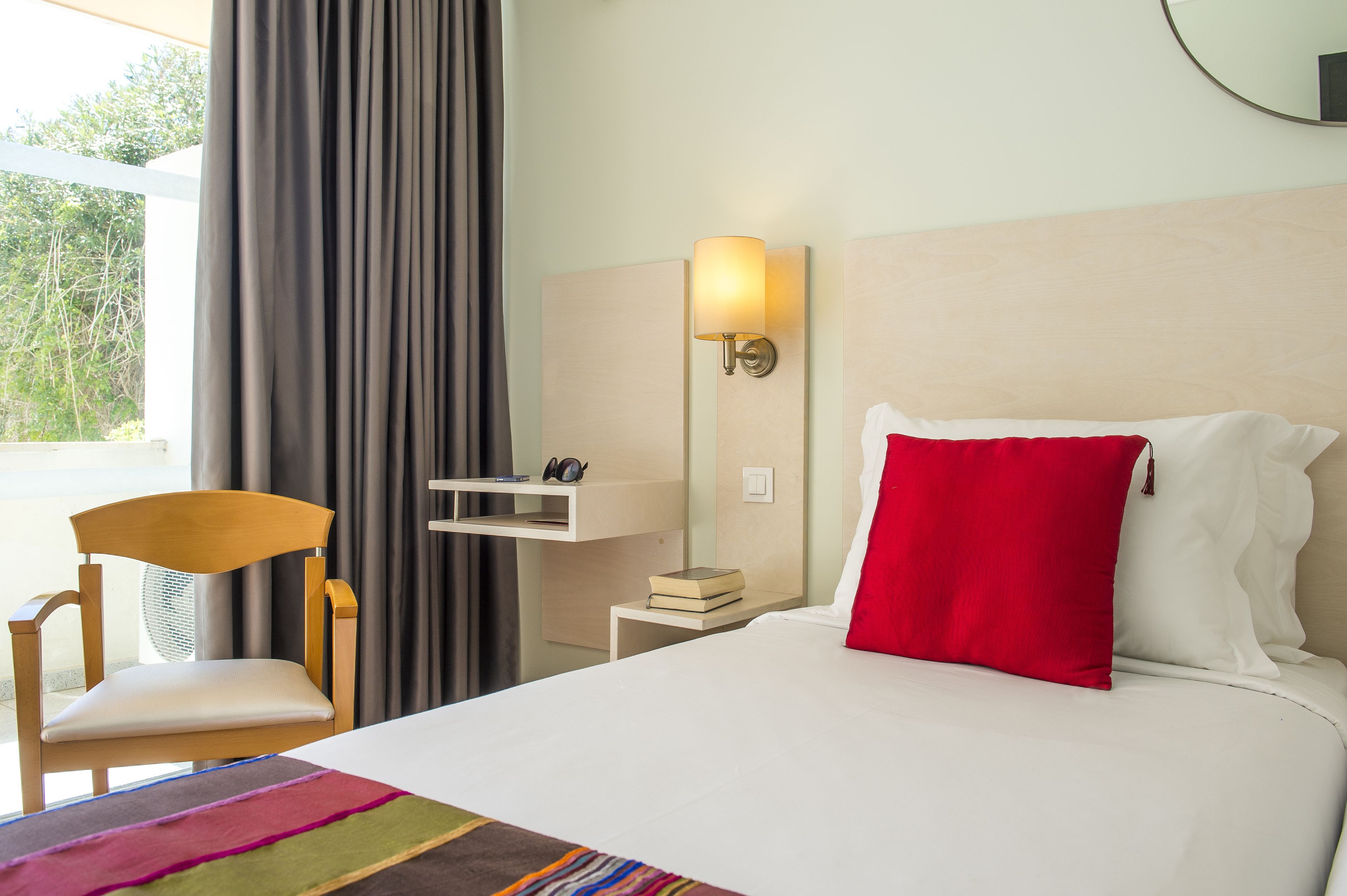 A hotel room with a bed, a chair, a bedside table with a lamp, and a view of the outdoors.