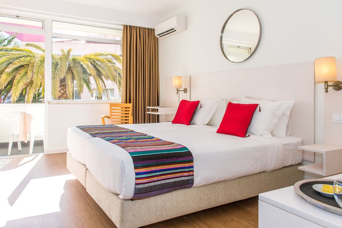 Habitación de hotel con una cama grande, un balcón con vista a palmeras, una ventana con cortinas y un espejo adosado a la pared.