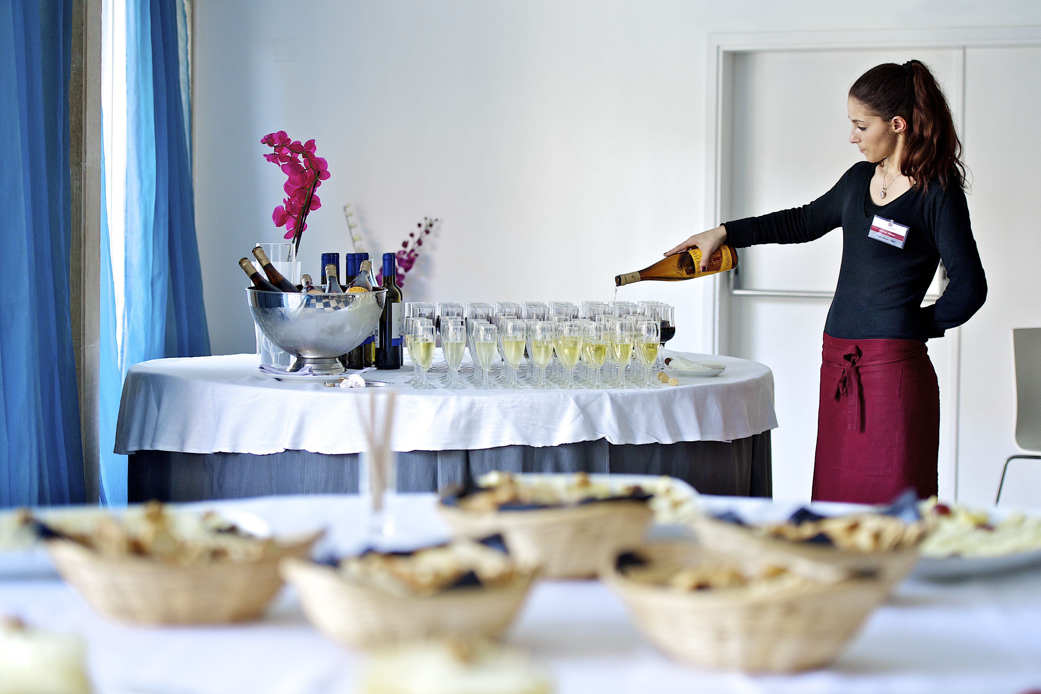 Une femme verse du champagne dans des verres sur une table de buffet où sont disposés divers plats et un vase décoratif.