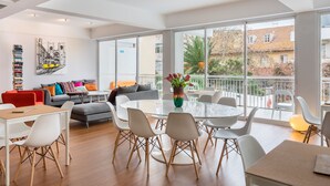 A spacious living area with a round glass table surrounded by chairs, a sofa, and a bookshelf.