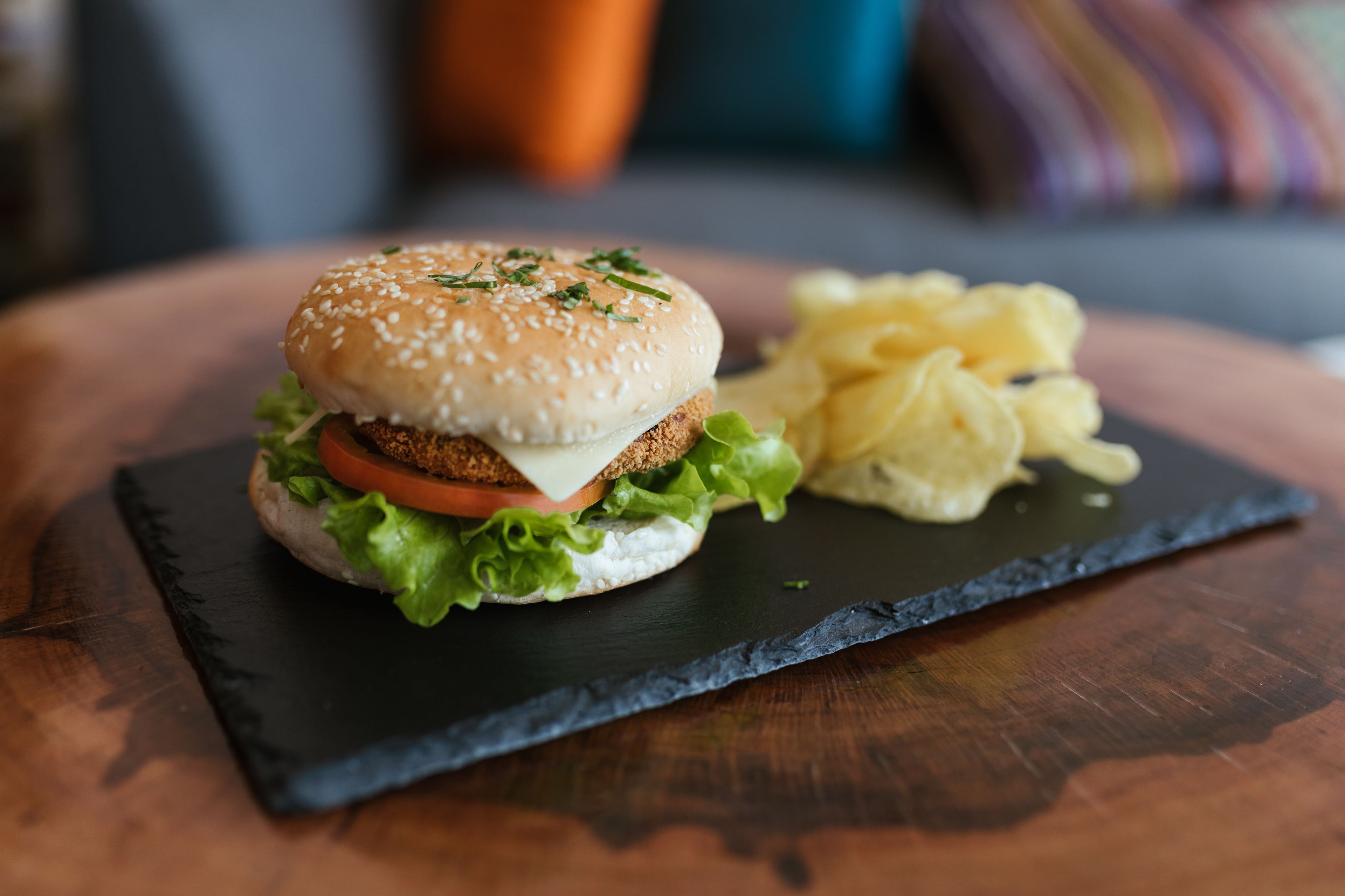 A burger with lettuce, tomato, and cheese on a sesame seed bun, served with potato chips on a slate board.