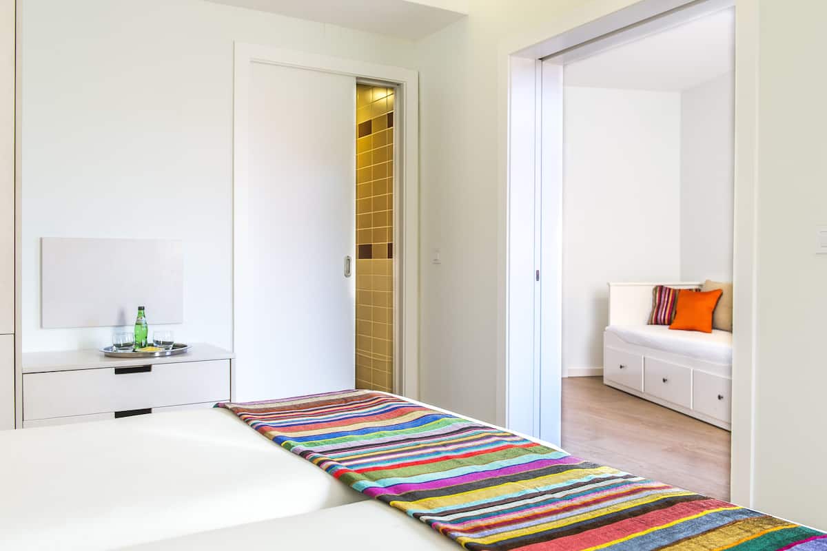 Una habitación de hotel moderna con una cama, una mesita de noche con una botella y vasos, una alfombra a rayas de colores, una pared blanca y una pequeña zona de estar con un cojín y una almohada.