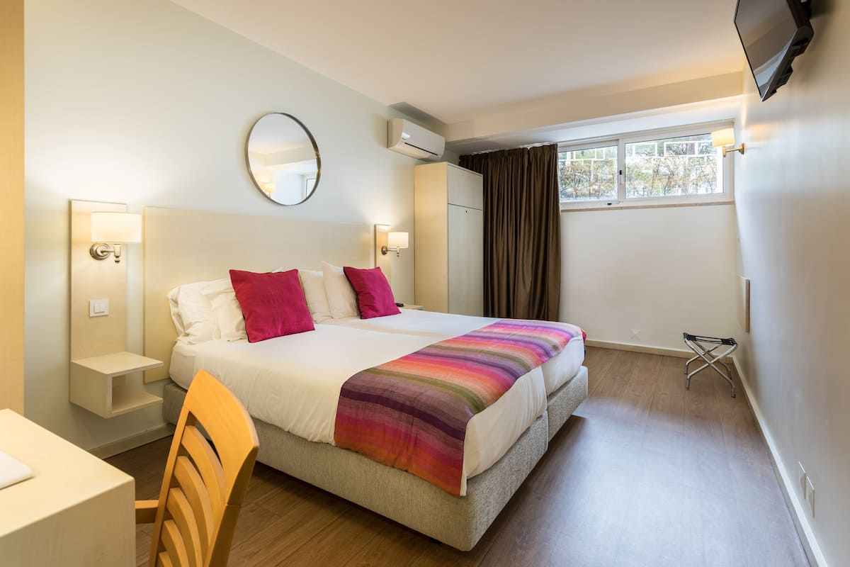 Habitación de hotel con cama, mesitas de noche, una silla, televisor y ventana con cortinas.
