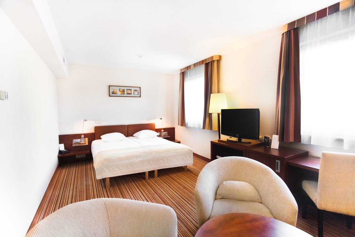 A hotel room with a bed, two chairs, a desk, a TV, and a lamp.