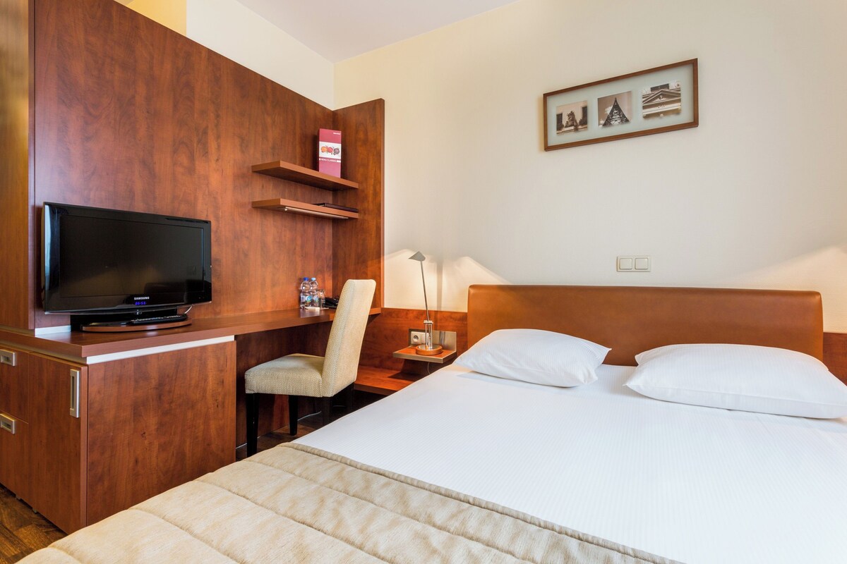 A hotel room with a bed, a TV on a wooden desk, a chair, a nightstand, and a wall-mounted shelf with a picture frame.