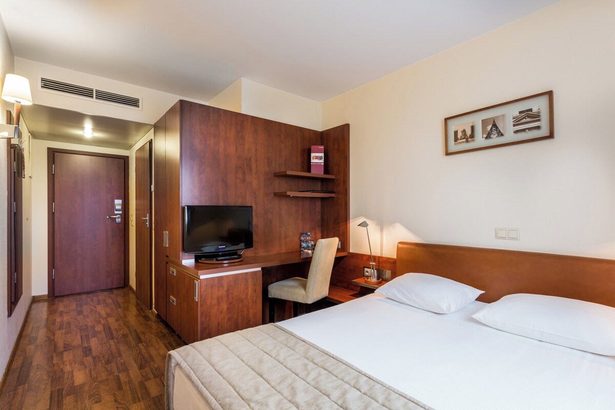 A hotel room with a bed, a desk with a television, a chair, a lamp, and a shelf with a book.